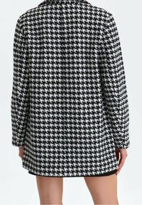 Svart- och vit hundtandsmönstrad kappa, gjord av texturerat tyg, med krage och långa ärmar, rak skärning och knälång fåll.