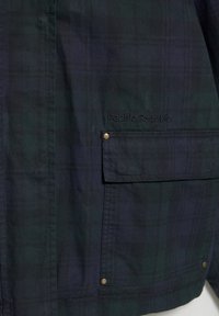 Kariertes Jackett in Dunkelgrün und Marineblau. Verfügt über eine Fronttasche mit Klappe, Messing-Druckknopf und gesticktem "Pacific Republic"-Logo. Glatte Textur.