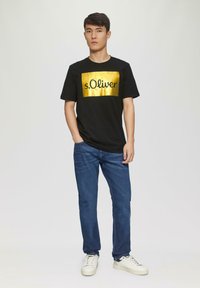 Zwart T-shirt met korte mouwen met een gouden rechthoekig logo, gecombineerd met blauwe spijkerbroek en witte sneakers, tegen een effen achtergrond.