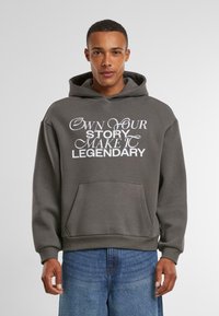 Sudadera gris con un gran diseño de texto impreso en blanco que dice "POSEE TU HISTORIA HAZLA LEGENDARIA". Bolsillo tipo canguro en la parte frontal, puños acanalados.