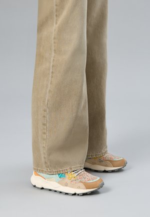 Bein in beigen, weit geschnittenen Hosen über Sneakern mit Blumenmuster und beigen, mint- und gelben Akzenten auf einem grauen Boden.