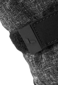 Schwarzer Stoff mit einer strukturierten Webart, versehen mit einem dunklen rechteckigen Etikett mit einem Logo. Die Nähte sind entlang der Kanten sichtbar.