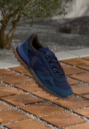 Navy blue athletic shoe with textured suede accents, lace-up design, and a rugged outsole. Positioned above a wooden tile and gravel surface.