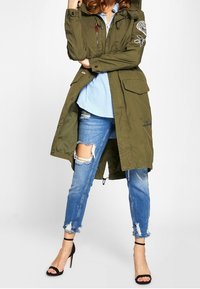 Olivgrön lång jacka med framfickor, huva och lappar, kombinerad med slitna blå jeans med fransade fållar och en ljusblå skjorta.