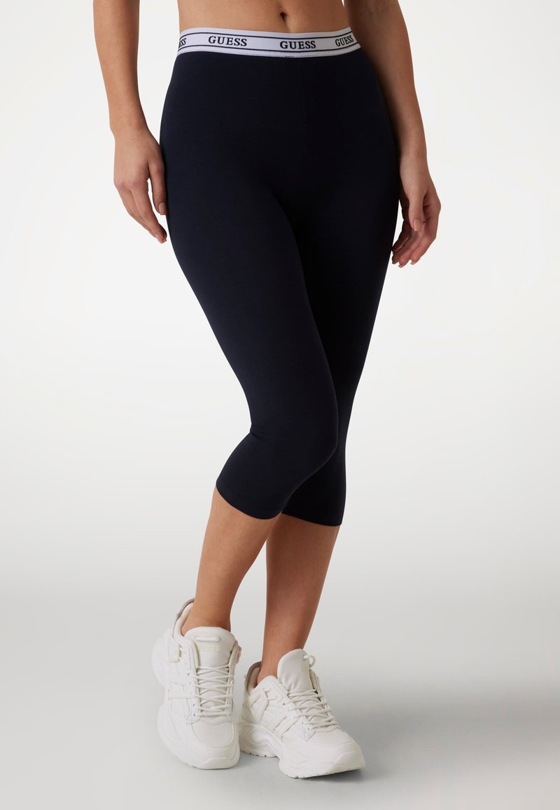 Leggings blu marino con un cinturino bianco con il logo GUESS, lunghezza a metà polpaccio, realizzati in tessuto elastico, abbinati a scarpe da ginnastica bianche.
