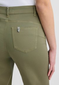Olivově zelené džíny s klasickým pětipocket designem, viditelným šitím a kovovým logem na zadní kapse.