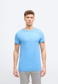 Νέος άνδρας που στέκεται με το πρόσωπο προς τα εμπρός, φορώντας ανοιχτό μπλε κοντομάνικο μπλουζάκι και γκρι σορτς, με φόντο μια απλή ανοιχτή επιφάνεια.