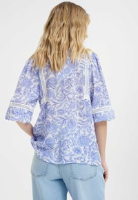 Bluza s plavim cvjetnim uzorkom i čipkastim detaljima, slobodnog kroja, tri četvrtine rukava, izrađena od teksturirane tkanine, u kombinaciji s svijetloplavim trapericama.