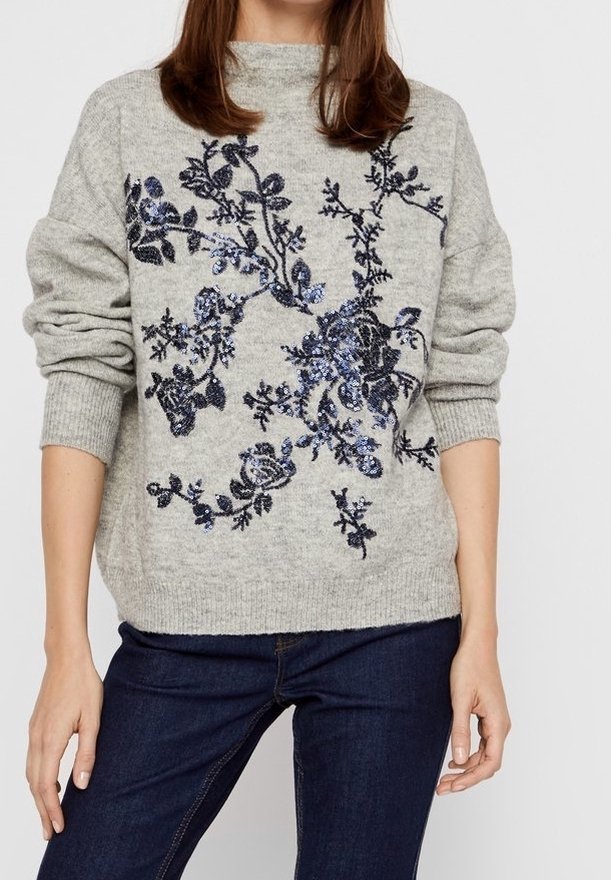 Femme portant un pull en maille gris clair avec une broderie florale bleue en sequins et un jean bleu foncé, debout devant un fond uni.