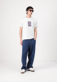 T-shirt en coton blanc avec un design graphique violet sur la poitrine, associé à un jean en denim bleu ample et des baskets blanches avec des accents beiges.