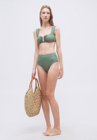 Γυναίκα με πράσινο μαγιό δύο τεμαχίων που κρατάει μεγάλη πλεκτή τσάντα παραλίας, όρθια ξυπόλυτη μπροστά σε απλό λευκό φόντο.