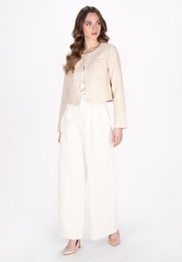 Giacca corta beige con design a quadri testurizzati, due tasche frontali e chiusura con bottoni, abbinata a pantaloni ampi bianchi e tacchi color nude.