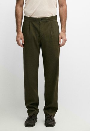 Homme portant un pantalon droit vert olive et des chaussures marron foncé à lacets, debout devant un fond clair uni.
