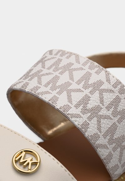Λευκό λουράκι σανδάλου με ανοιχτό καφέ μοτίβο "MK" και χρυσό κυκλικό λογότυπο MK σε φόντο μπεζ δέρματος.