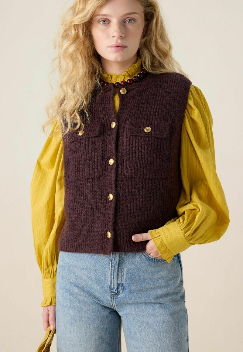 Bordeaux wollen vest met twee voorzakken en gouden knopen, gelaagd over een gele blouse met ruches aan de manchetten, gecombineerd met blauwe jeans.