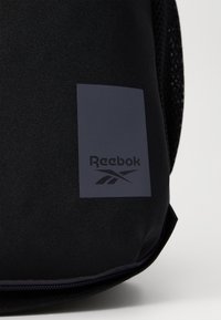 Melnā mugursoma, kas izgatavota no izturīga auduma, ar taisnstūra formas logo plātni tumši zblue, uz kuras ir teksts "Reebok" un izteiksmīgs logo dizains.