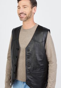 Schwarze Lederweste mit glatter Textur, ausgestattet mit V-Ausschnitt, Knopfverschluss und zwei Seitentaschen. Über einem beigen Pullover getragen.