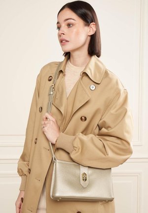Jeune femme portant un trench-coat beige et tenant un sac à main doré métallique sur l'épaule, debout à l'intérieur contre un mur crème.