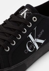 Černé plátěné tenisky s bílým logem "CK" a textem "Calvin Klein Jeans". Kovové očka a texturovaná černá gumová podrážka. Jednoduchý design.