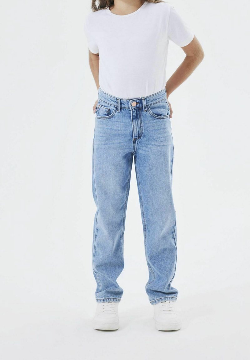 Enfant portant un jean taille haute bleu clair, un t-shirt blanc et des baskets blanches, debout les mains derrière le dos sur un fond blanc.