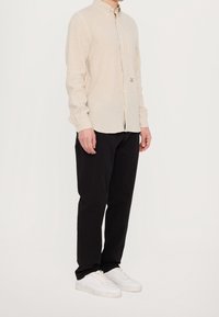 Uomo che indossa una camicia beige con bottoni, pantaloni neri e sneakers bianche, in piedi contro uno sfondo bianco uniforme.