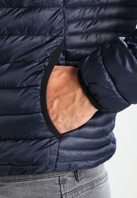 Marineblå puffy jakke med lodret quiltet mønster, med en sidelomme og sorte kanter ved åbningen. Glat, letvægtsstof.