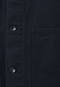 Camisa de algodón negra de botones con un bolsillo en el pecho y un discreto logo de Nike bordado en el bolsillo. Botones plateados visibles.
