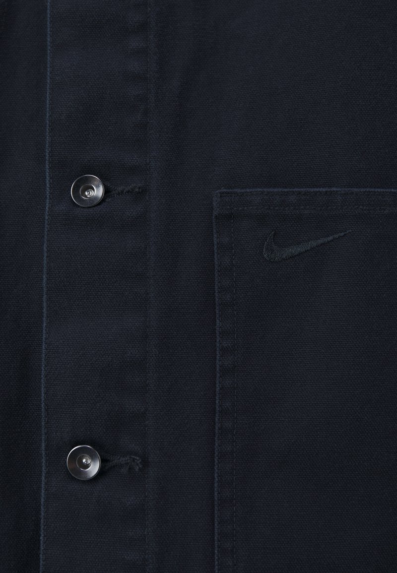 Camisa de algodón negra de botones con un bolsillo en el pecho y un discreto logo de Nike bordado en el bolsillo. Botones plateados visibles.