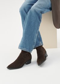 Stivaletti in suede marrone con punta affusolata e tacco basso, abbinati a jeans blu, posizionati su uno sfondo neutro.