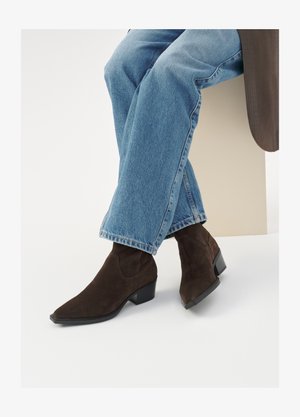 Brown suede ankle boots with a pointed toe and low block heel, paired with blue denim jeans, sitting against a neutral background.