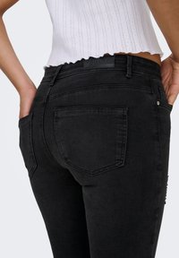 Black denim jeans with a fitted design, featuring two back pockets and minimal stitching detail. The waistband includes a small brand tag.