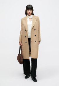 Beiger Wollmantel mit doppelseitigem Design und schwarzen Knöpfen, getragen über einem weißen Oberteil und schwarzen Hosen; accessoriert mit einer braunen Tasche.