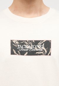 T-shirt blanc avec un patch rectangulaire noir et beige sur la poitrine portant l'inscription "JACK & JONES Remote Community" en texte blanc.