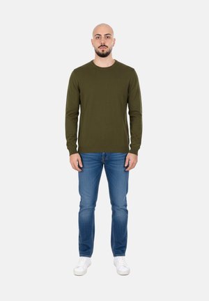 Uomo con barba che indossa una camicia verde oliva a maniche lunghe, jeans blu e scarpe da ginnastica bianche, in piedi davanti a uno sfondo chiaro e uniforme.