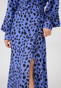 Robe bleue avec un motif à pois noirs, dotée de manches longues, d'une taille élastique et d'une fente latérale, fabriquée en tissu lisse. Détail froncé sur le côté.