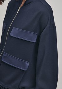 Námořnická bombardovací bunda vyrobená z hladké látky. Má dvě velké našité kapsy, zipový uzávěr a stojáček na krku.