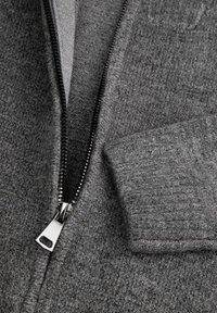 Zbliżenie na szary dziergany sweter z częściowo otwartym srebrnym zamkiem błyskawicznym i widocznym prążkowanym ściągaczem na rękawie.