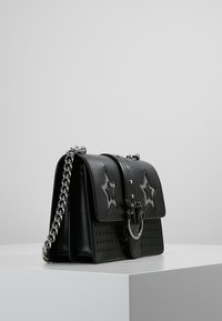 Borsa a tracolla in pelle nera con intagli a stella, tracolla a catena e un fermaglio metallico circolare. Presenta borchie decorative e una superficie liscia.