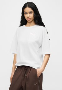 Νεαρή γυναίκα με μακριά σκούρα μαλλιά, που φοράει λευκό μπλουζάκι Puma και καφέ παντελόνι με κορδόνι και λογότυπο Puma, στέκεται με το ένα χέρι στην τσέπη.