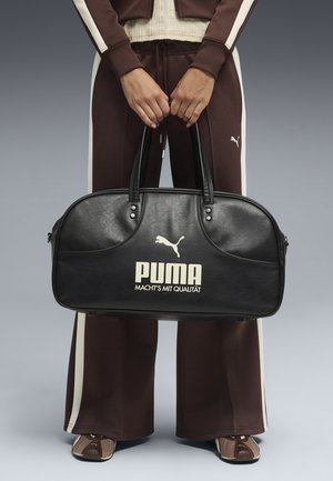 Osoba w brązowym dresie Puma trzymająca czarną torbę sportową Puma z białym logo i napisem, stojąca na tle szarego tła.