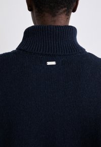 Ναυτικά μπλε πλεκτό πουλόβερ με ψηλό ριμπ γιακά και ασημένια ετικέτα λεπτομέρεια στην πλάτη. Το ύφασμα έχει απαλό, υφασμένο εμφάνιση.