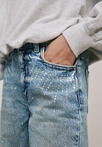 Blugi din denim cu spălare albastru deschis, cu paiete transparente aranjate într-un model punctat și un buzunar frontal vizibil.