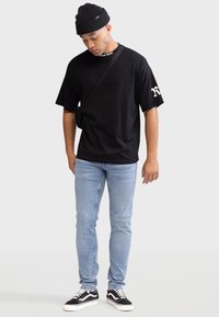 Schwarzes Oversized-T-Shirt mit weißem Grafikaufdruck auf dem Ärmel, blaue Skinny Jeans, schwarze Sneakers mit weißer Sohle und eine schwarze Strickmütze.
