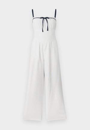 Valkoinen pellavainen haalari, jossa on leveät lahkeet, rypytetty yläosa ja laivastonsiniset hihnat, joissa on solmukuvio keskellä.