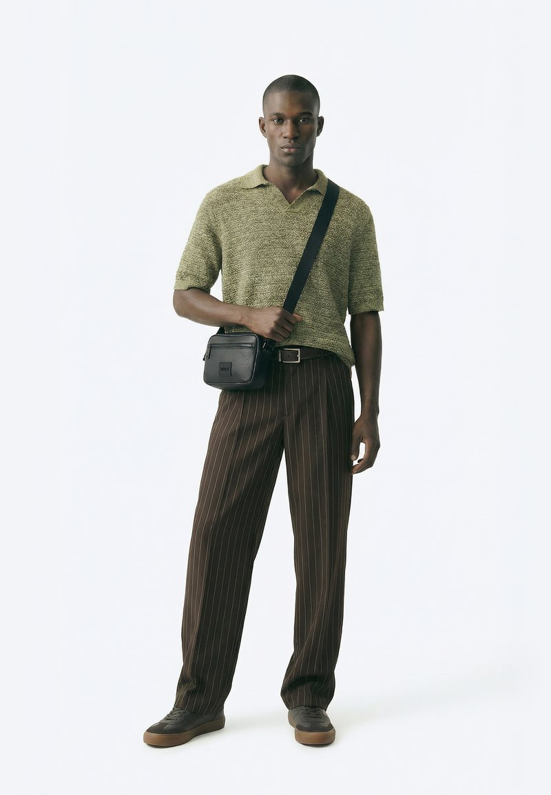 Zöld kötött galléros pólót, barna csíkos nadrágot, szürke tornacipőt és fekete keresztpántos táskát viselő férfi fehér háttér előtt áll.