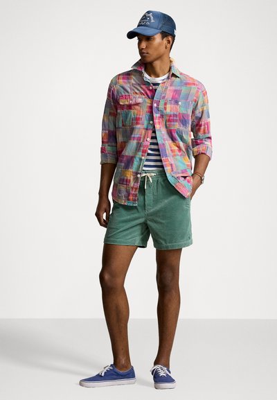 Chemise à carreaux colorée avec deux poches avant, t-shirt rayé, shorts en velours côtelé vert et chaussures en toile bleues. Casquette avec détail de logo.