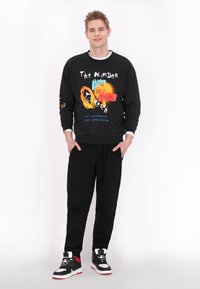 Hanorac negru cu text grafic colorat, mâneci lungi și manșete cu jaretă, asortat cu pantaloni negri și adidași cu vârf înalt negri și roșii.
