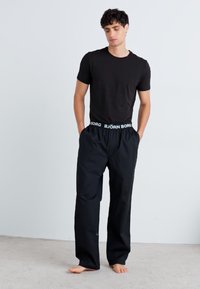 Camiseta preta e calças relaxadas pretas com uma cintura elástica branca com o texto "BJÖRN BORG", em pé sobre um chão cinza claro.
