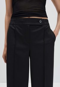 Vrouw in hoge taille zwarte wijde broek met een voorzichtige plooi en een zwarte knoopdetail, gecombineerd met een zwart crop top.