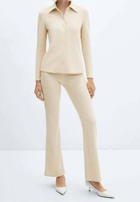 Ensemble beige texturé deux pièces avec un haut à manches longues, col à boutons et un pantalon évasé assorti, porté avec des talons blancs pointus.